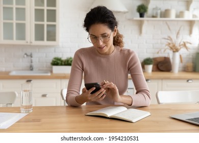 Confident young hispanic female work from home sit at desk search information online using internet resources via phone. Young woman freelancer texting client set up business meeting at suitable time