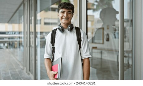 Confident young hispanic teenager, a joyful student wearing headphones, happily holding his university books, flashing a handsome, cheerful smile while casually standing on the lively city campus. - Powered by Shutterstock