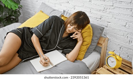 Confident young hispanic woman blissfully writing notes during a relaxing morning phone conversation, comfortably sprawled on her bedroom bed, her beautiful face lighting up her cozy apartment.