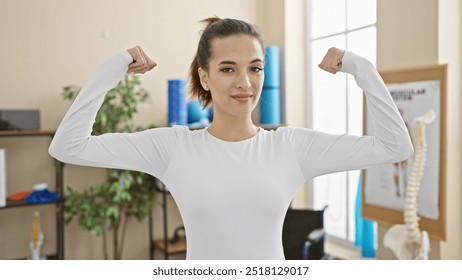 Confident young hispanic woman poses in a bright gym, wearing athletic attire and flexing muscles with equipment in the background. - Powered by Shutterstock