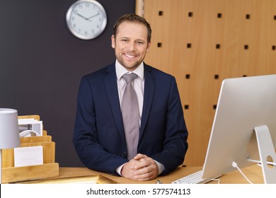 Confident Young Hotel Manager With A Welcoming Smile Standing Waiting To Greet Visitors At The Front Desk Of A Luxury Hotel