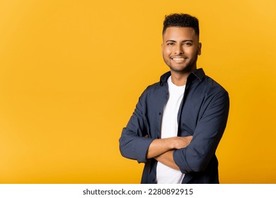 Confident young indian man standing in confident pose with arms crossed, looking at camera, copy space for advertising new promotion, arab guy standing isolated on yellow background.