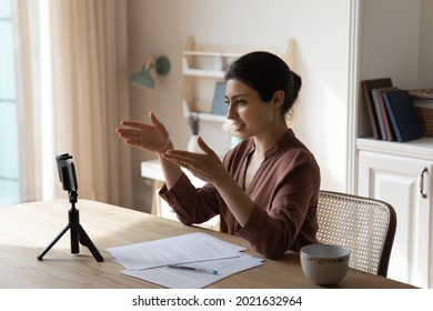 Confident Young Indian Woman Sit At Desk At Home Talk Speak On Video Call On Cellphone Record Webinar. Millennial Ethnic Female Coach Or Speaker Have Webcam Virtual Digital Event Shoot On Smartphone.