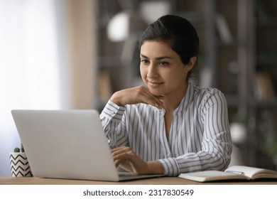 Confident young indian woman worker sit at office table by laptop search data browse corporate network for business news. Smiling female freelancer read document online correspond with client in chat - Powered by Shutterstock