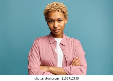 Confident young lady in casual pink clothes dyed in blonde short curls and nose ring standing against blue background with bossy look, posing with folded arms, looking skeptical with smirking smile
