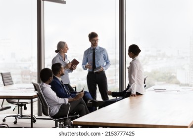 Confident Young Male Boss Talking To Diverse Team Of Managers, Discussing Work Issues On Corporate Meeting, Briefing. Business Teacher Training Students At Big Panoramic Window In Open Office Space