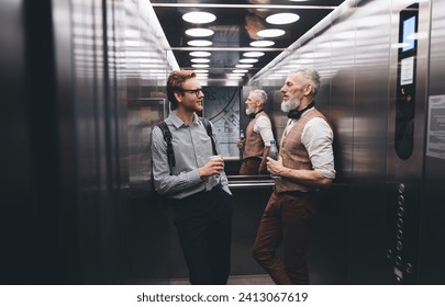 Confident young male entrepreneur and optimistic elderly businessman in formal wears standing and chatting in elevator in modern corridor with glass wall
