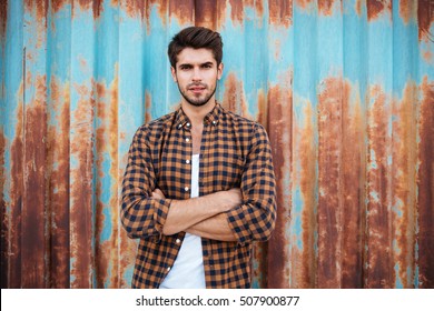 Confident young man in checkered shirt standing with arms crossed over blue metal background with rust - Powered by Shutterstock