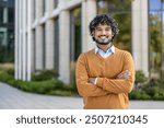 Confident young man with curly hair smiling outside modern office building. Wearing orange sweater with arms crossed, expressing positivity and professionalism. Reflects successful business outlook