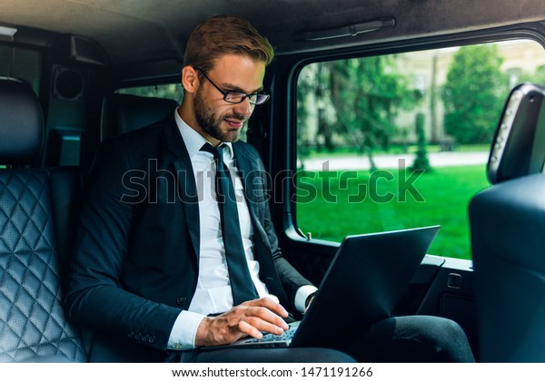 Confident young man\
in full suit working using laptop while sitting in the car being\
fully concentrate on\
work