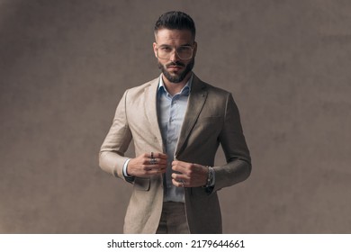 Confident Young Man With Glasses Closing Elegant Suit And Being Cool In Front Of Beige Background In Studio