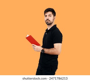 Confident Young Man Holding a Red Book Against an Orange Background. Boy with Book Wearing black shirt and black jeans. Young Male Student looking towards camera - Powered by Shutterstock