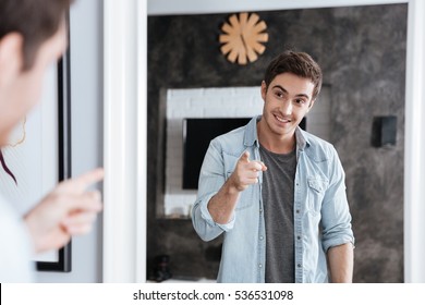 Confident young man pointing finger at his mirror reflection while standing at home