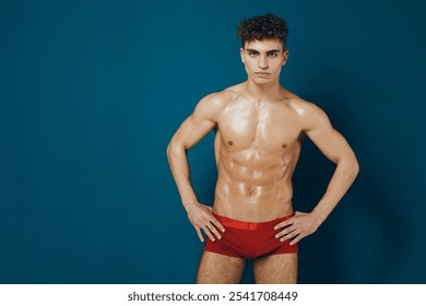 Confident young man posing shirtless in red underwear against a vibrant blue background, showcasing fitness and body positivity. - Powered by Shutterstock