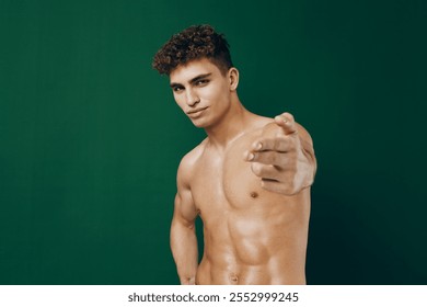 Confident young man posing shirtless against a green background, showcasing his fitness and strength. Strong posture with an inviting gesture. - Powered by Shutterstock