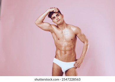 Confident young man posing in white underwear against a soft pink background, showcasing a fit physique and playful demeanor. Fitness and fashion concept. - Powered by Shutterstock