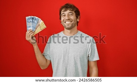Confident young man smiling while holding heap of hungary forint banknotes, isolated on red background, feeling rich and enjoying financial success
