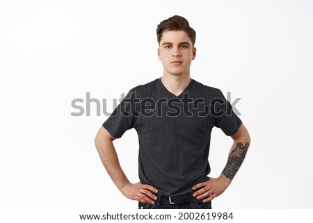 Confident young man standing with hands on waist, smiling and looking determined, ready for work, waiting for task, posing in t-shirt and jeans, white background