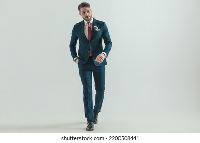 Confident Young Man Wearing Elegant Suit With Red Tie And Handkerchief, Holding Hand In Pocket And Walking On Grey Background In Studio