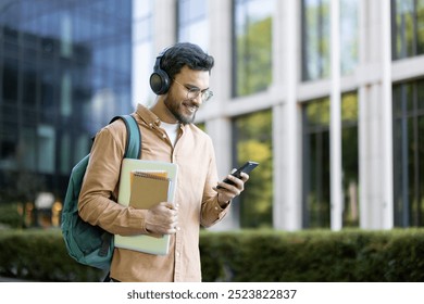 Confident young man wearing headphones walking in city holding backpack and notepad, checking smartphone. Represents lifestyle, technology, education, and casual communication in urban environment. - Powered by Shutterstock