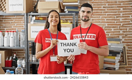 Confident young man and woman volunteers, standing in unity at charity center, holding a heartfelt 'thank you' paper banner in spotlight of altruism.