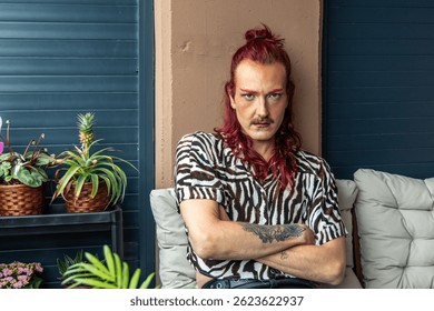 Confident young non-binary person with pink hair and makeup sitting on balcony sofa with arms crossed, surrounded by plants, celebrating self-acceptance, pride, and body positivity
