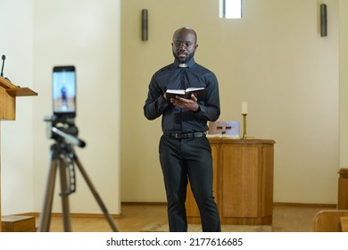 Confident young pastor of evangelical church with open Holy Bible standing in front of smartphone camera and pronouncing sermon - Shutterstock ID 2177616685