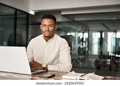 Confident young professional African American business man looking at camera sitting at work desk using computer. Portrait of male company employee or entrepreneur with laptop in office, copy space. - Powered by Shutterstock