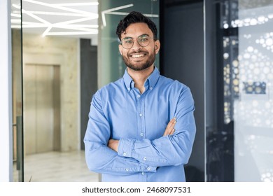 Confident young professional wearing glasses and blue shirt standing in a modern office environment with arms crossed and a smile. - Powered by Shutterstock