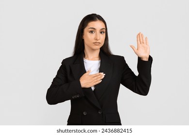 Confident young professional woman in black suit takes an oath, placing one hand on chest and other raised, standing against light gray backdrop - Powered by Shutterstock