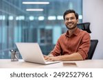 Confident young professional working on laptop in modern office. Smiling and looking at camera, embodies productivity and job satisfaction in a professional setting.