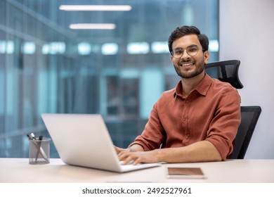 Confident young professional working on laptop in modern office. Smiling and looking at camera, embodies productivity and job satisfaction in a professional setting. - Powered by Shutterstock