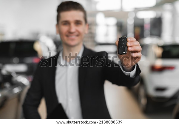 Confident
young salesman showing car key at camera in modern dealership,
selective focus. Millennial auto dealer selling automobiles at
showroom. Vehicle local distribution
concept
