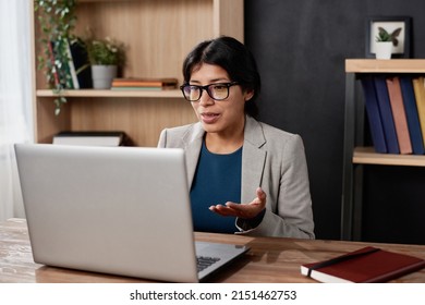 Confident Young Spanish Teacher In Jacket Sitting At Table In Office And Using Laptop While Talking To Student Via Videoconferencing App