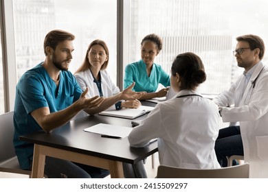 Confident young surgeon man in blue medic uniform talking to multiethnic colleagues in hospital meeting room, speaking to listening diverse doctors at table, offering ideas for clinic teamwork - Powered by Shutterstock