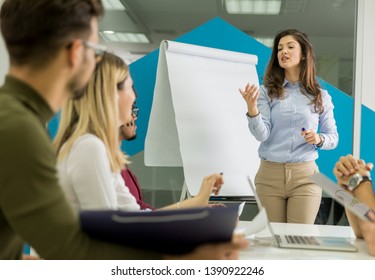 Confident Young Team Leader Giving A Presentation To A Group Of Young Colleagues As They Sit Grouped By The Flip Chart In The Small Startup Office