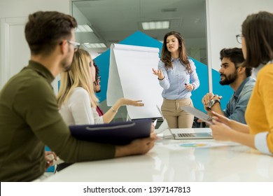 Confident Young Team Leader Giving A Presentation To A Group Of Young Colleagues As They Sit Grouped By The Flip Chart In The Small Startup Office