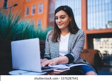 Confident Young Woman In Casual Outfit And Glasses Typing On Laptop Keyboard While Sitting On Bench Outside Building On Modern City Street