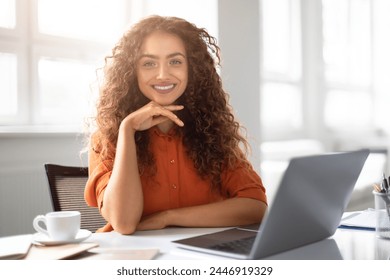 A confident young woman with curly hair smiling at the camera while working on her laptop in a bright office setting - Powered by Shutterstock