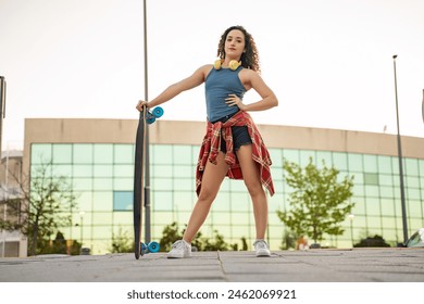 A confident young woman with curly hair holding a skateboard, wearing headphones and a blue tank top, standing in an urban park at sunset.  - Powered by Shutterstock