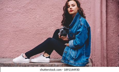 Confident young woman with curly long hair wearing denim jacket and black trousers sitting on stone bench with smartphone and holding retro photo camera - Powered by Shutterstock