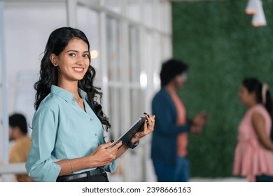 confident young woman employee standing at office by looking camera with tab in hand - concept of businesswoman, startup entrepreneur and career growth