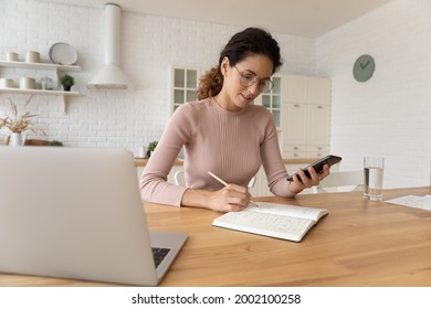 Confident young woman in eyeglasses sitting at table with computer, web surfing information using mobile phone app. writing notes in copybook, studying preparing for exams or managing affairs. - Powered by Shutterstock