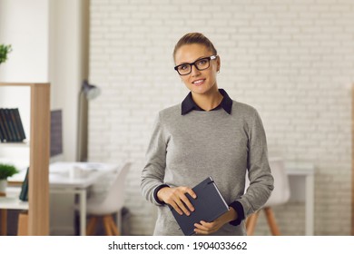 Confident young woman in glasses and formal wear standing in office, holding notebook and smiling. Portrait of professional business advisor, smart student, school teacher or faculty member at work