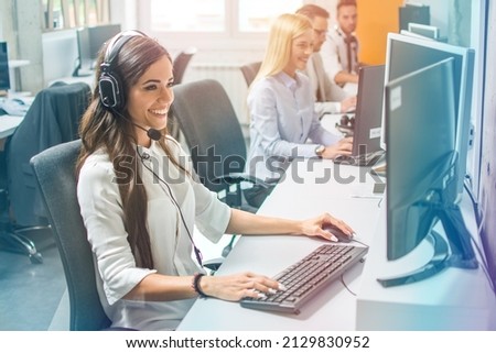 Confident young woman in headset, sitting in office, working as operator of call center or support service with her colleagues.