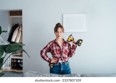 Confident young woman in protective glasses with electric screwdriver in hand in after she put picture on the wall at her home. Housekeeping work. Doing repair herself. Home decorating. DIY concept.