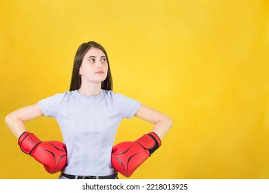 Confident Young Woman With Red Boxing Gloves Stands With Hands On Hips Looking Aside. Portrait Of Strong And Determined Girl Isolated On Yellow Background. Self Defence And Leadership Concept