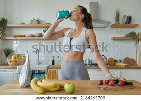 Confident young woman in sports clothing drinking protein cocktail while standing at the kitchen