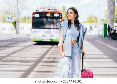 Confident young woman stands at city bus stop with suitcase and laptop. Dressed in casual attire, she exudes modern traveler vibe. - Powered by Shutterstock