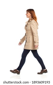 Confident Young Woman Is Walking In Beige Coat, Jeans And Brown Shoes. Side View. Full Length Studio Shot Isolated On White.